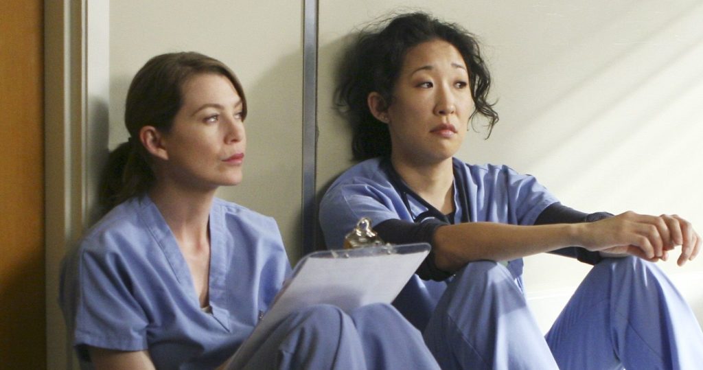 Meredith Grey (Ellen Pompeo) and Cristina Yang (Sandra Oh) sitting against a wall in the hospital in 'Grey's Anatomy'