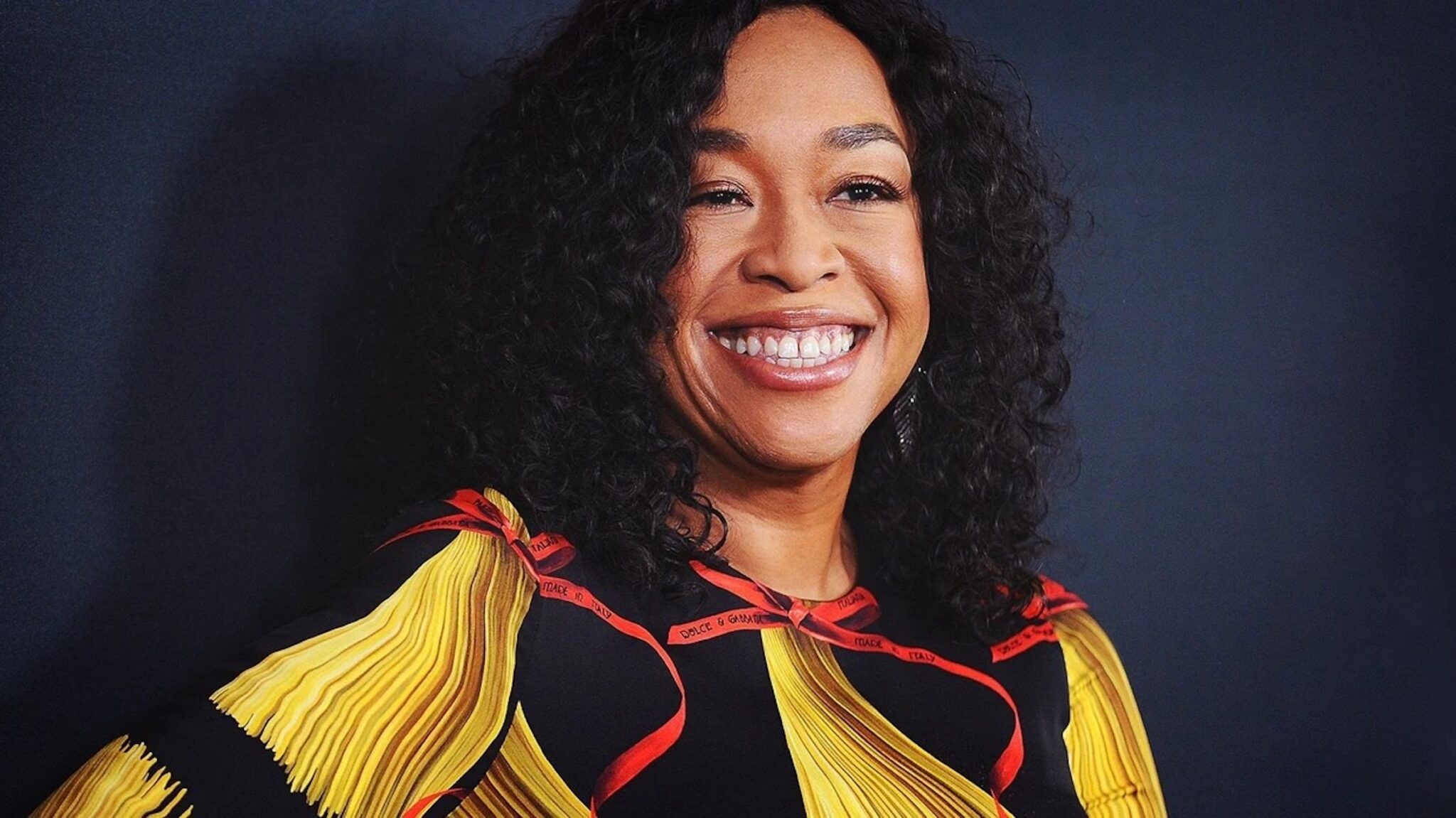 Shonda Rhimes standing against a dark blue background in a yellow and black dress, 5 Trademarks of Shonda Rhimes